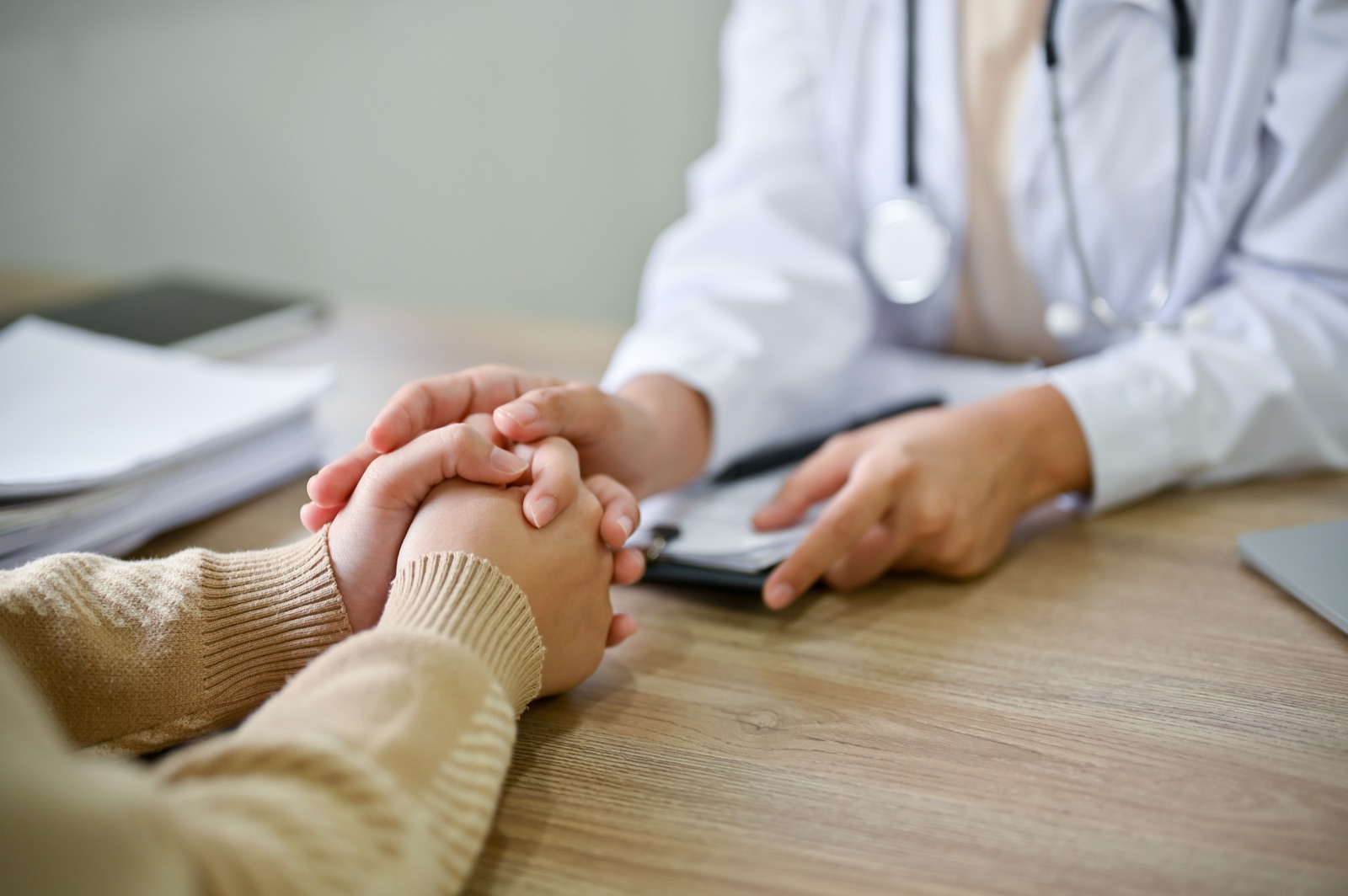 A doctor reaches across a desk to hold a patient's hands during a consultation